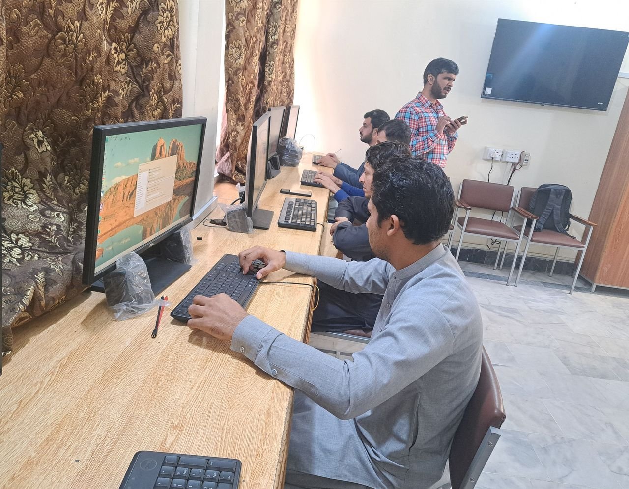 A trainer stands in the foreground while participants work at computer workstations behind him.