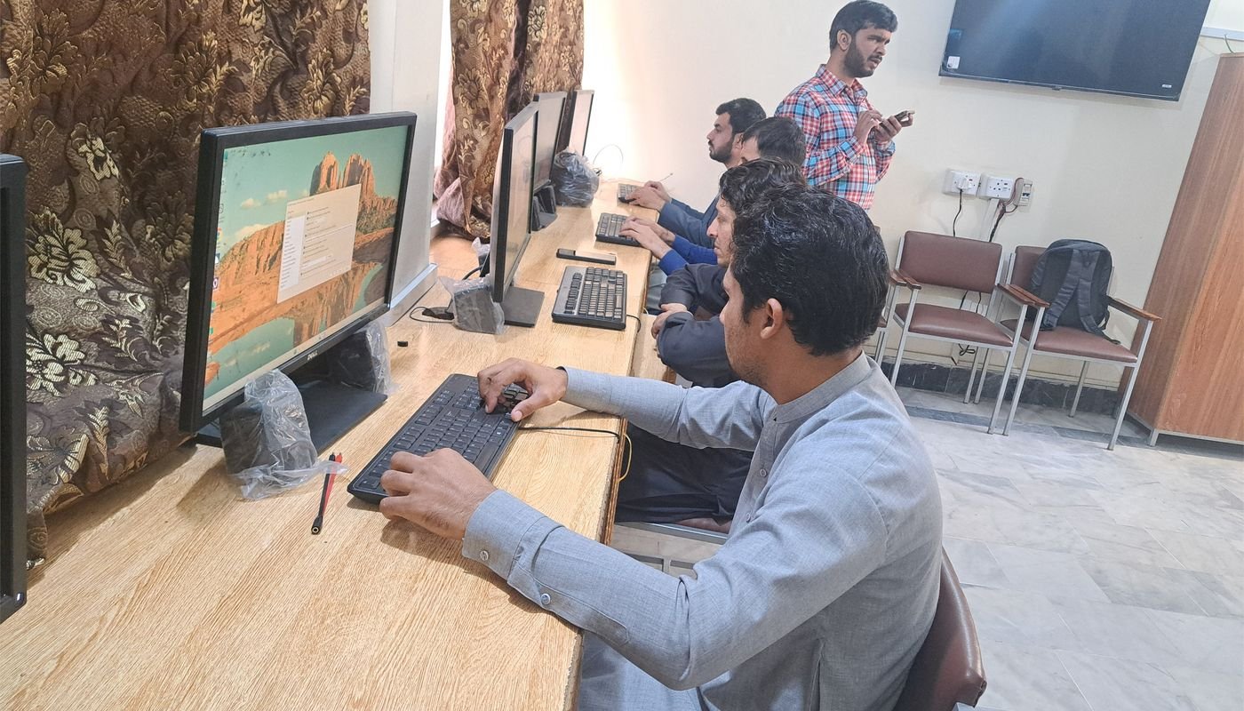 A trainer stands in the foreground while participants work at computer workstations behind him.