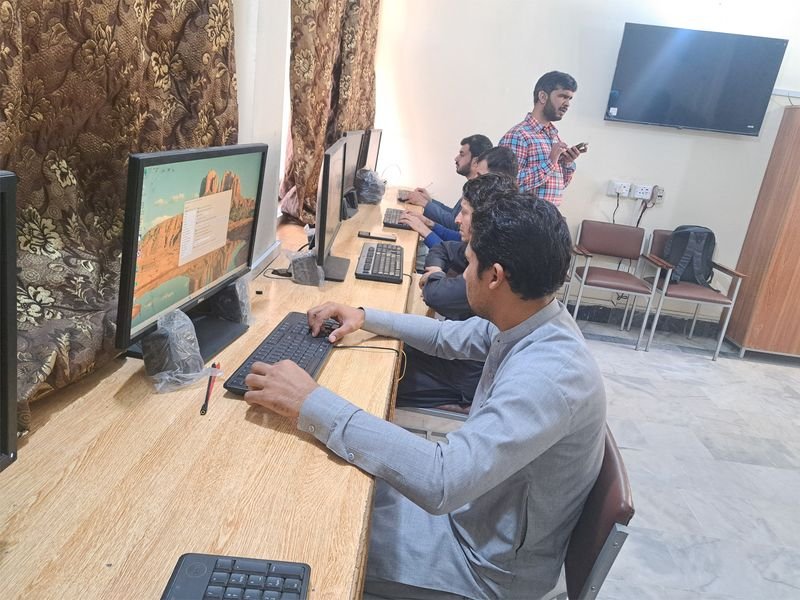 A trainer stands in the foreground while participants work at computer workstations behind him.